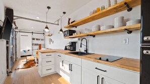 A modern kitchen with white cabinets, a wooden countertop, and black appliances. There are hanging lights and shelves above the counter. - Cowboy Shipping Container With Private Hot Tub (Columbia Falls)