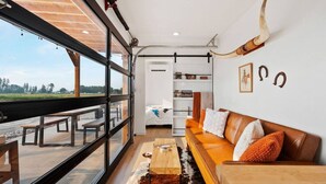 A modern living room with a large window, a leather sofa, a wooden coffee table, and a cowhide rug. - Cowboy Shipping Container With Private Hot Tub (Columbia Falls)