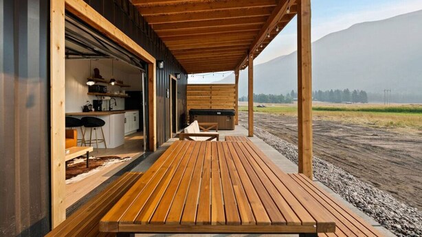 A wooden deck with a table and chairs, a kitchen area with bar stools, and a view of a mountainous landscape. - Cowboy Shipping Container With Private Hot Tub (Columbia Falls)