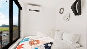 A bedroom with a large window, a bed with a colorful quilt, a white air conditioning unit, and a small table with a chair. - Cowboy Shipping Container With Private Hot Tub (Columbia Falls)