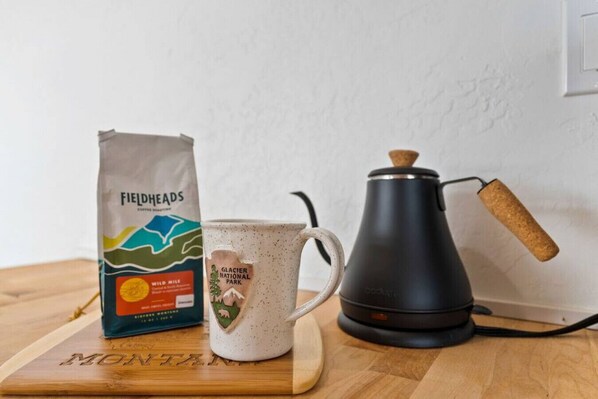 A bag of Fieldheads coffee, a white mug with a Glacier National Park design, and a black coffee press on a wooden surface.