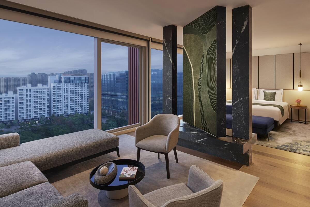 A modern hotel room with a large window offering a city view, a comfortable seating area, a bed with a view, and a decorative wall panel.