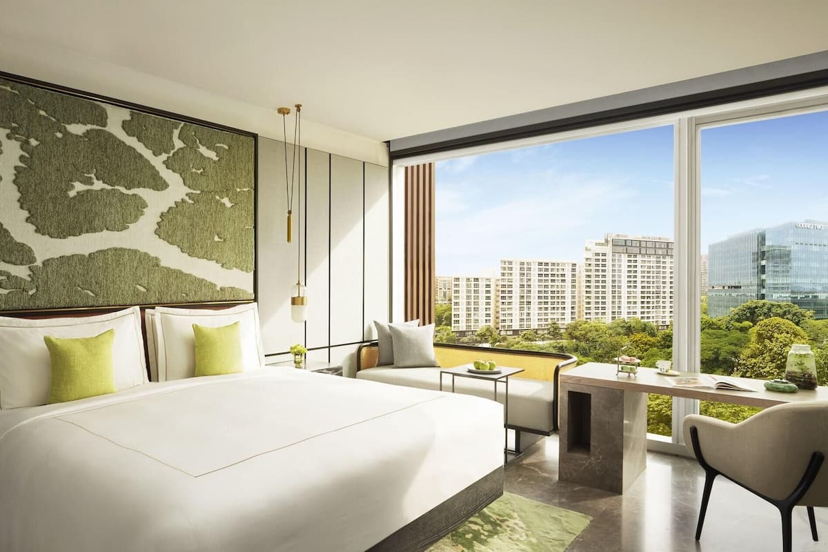 A modern hotel room with a large bed, a seating area, a dining table, and a view of the cityscape.