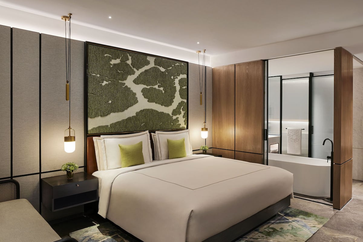 A modern hotel room with a large bed, a nightstand, a wall-mounted lamp, and a bathroom with a bathtub and mirror.