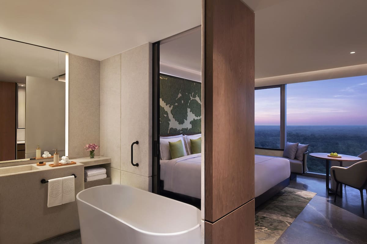 A modern hotel room with a large bathtub, a bed with pillows, a seating area with a table, and a view of the landscape through large windows.