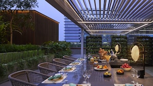 Ein Essbereich im Freien mit einem langen Tisch, gedeckt für eine Mahlzeit, umgeben von Korbstühlen und einem überdachten Pergola.