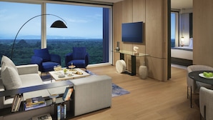 Ein modernes Wohnzimmer mit großem Fenster, zwei blauen Sessel, einem grauen Sofa, einem Couchtisch mit Büchern und einem flachbildfernseher an der Wand.