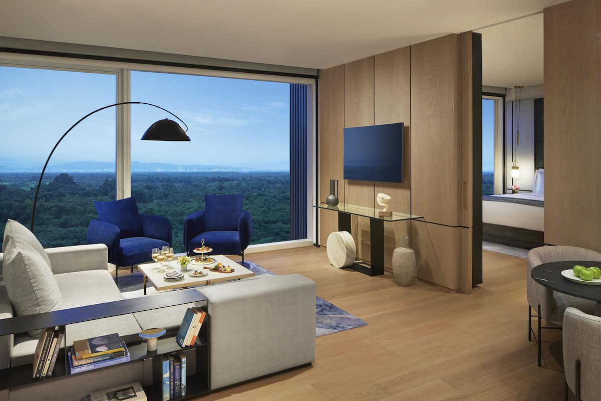 A modern living room with a large window, two blue armchairs, a grey sofa, a coffee table with books, and a flat-screen TV mounted on the wall.