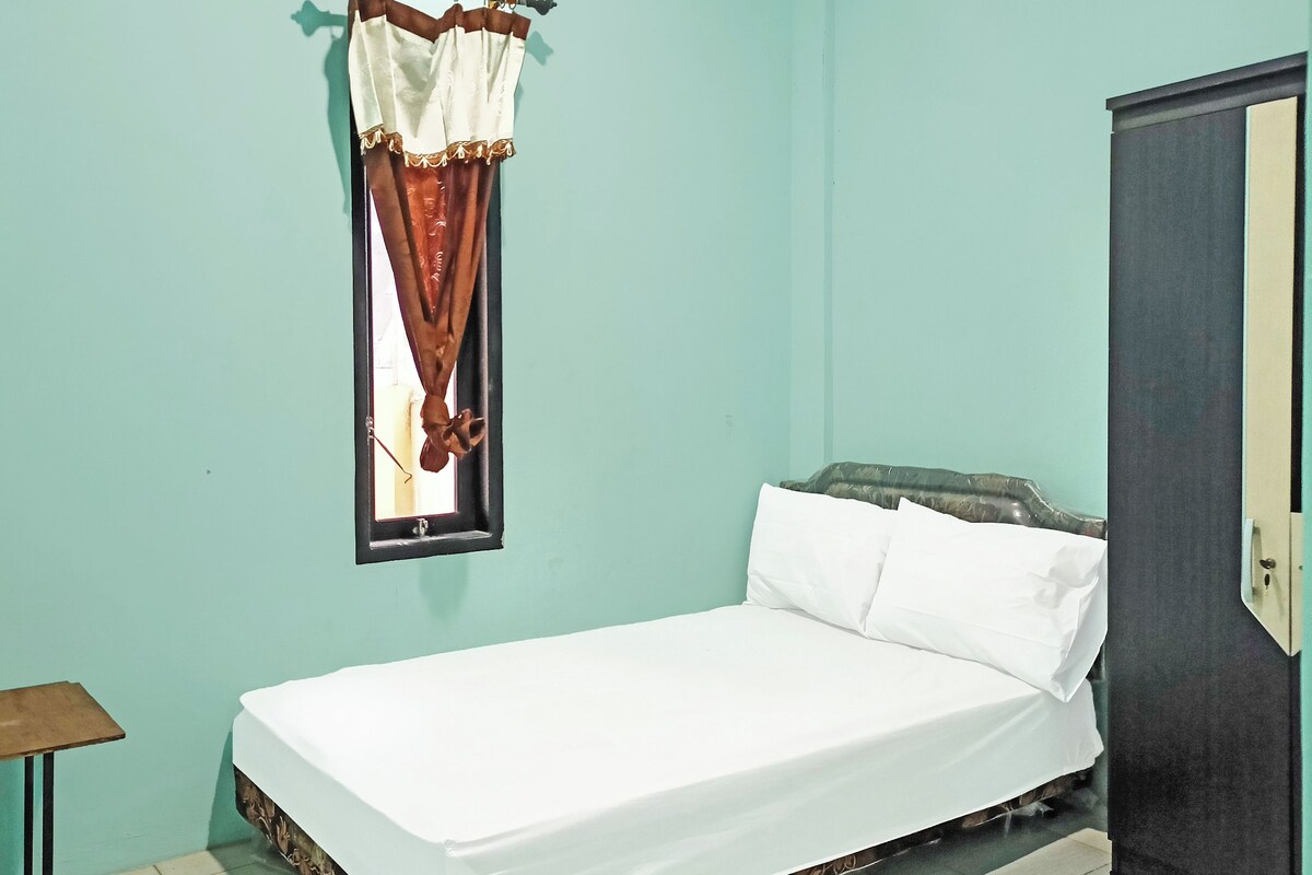 A single bed with white bedding, a window with brown curtains, a small wooden table, and a door.