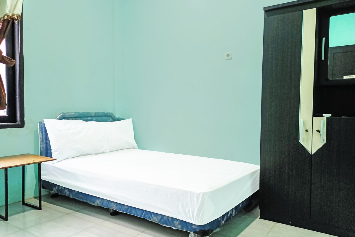 A single bed with a white mattress and blue headboard, a small wooden table, and a dark-colored wardrobe in a room with light blue walls.
