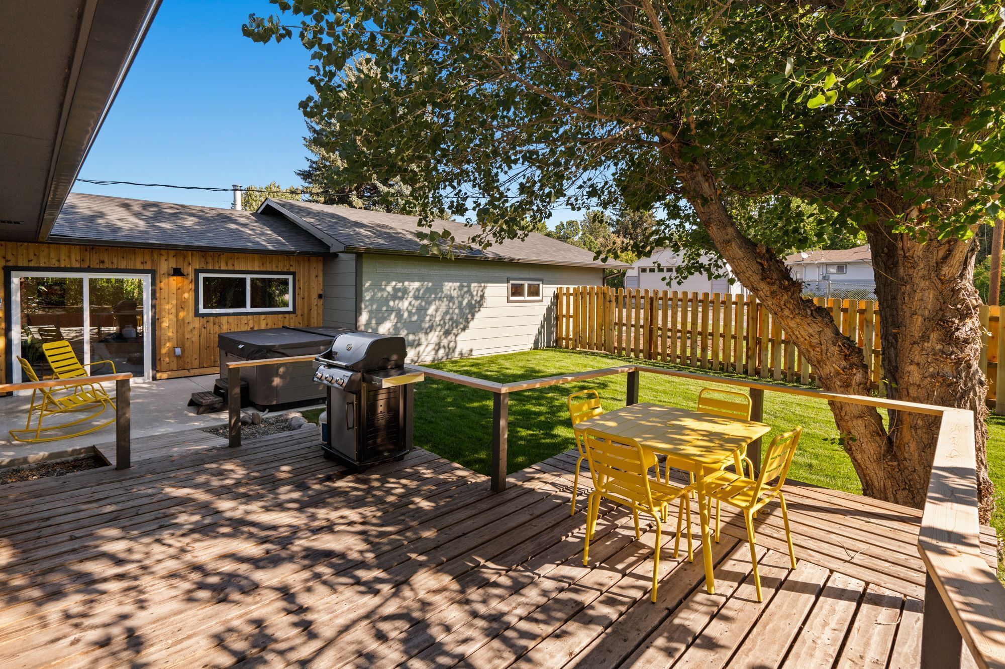 A backyard with a wooden deck, a barbecue grill, a table with yellow chairs, and a small shed.
