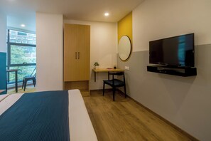 Habitación de hotel con cama, escritorio con silla, televisor y ventana con vista a edificios y vegetación.