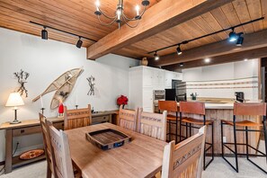 A dining area with a wooden table and chairs, a chandelier, and a kitchen in the background. - Enclave 208 | Snowmass Ski-In/Ski-Out Condo w/Shared Pool/Hot Tub/WiFi (Snowmass Village)