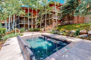 A pool area with a clear pool, surrounded by trees and a building with multiple balconies. - Enclave 208 | Snowmass Ski-In/Ski-Out Condo w/Shared Pool/Hot Tub/WiFi (Snowmass Village)