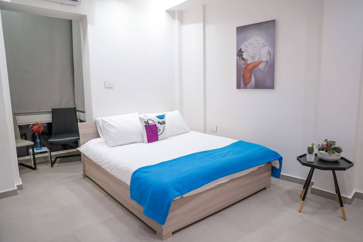 A modern bedroom with a bed, a chair, a small table with a plant, and a painting on the wall.