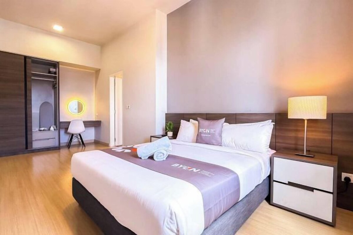 A modern hotel room with a large bed, bedside tables, a nightstand, a mirror, and a wardrobe.