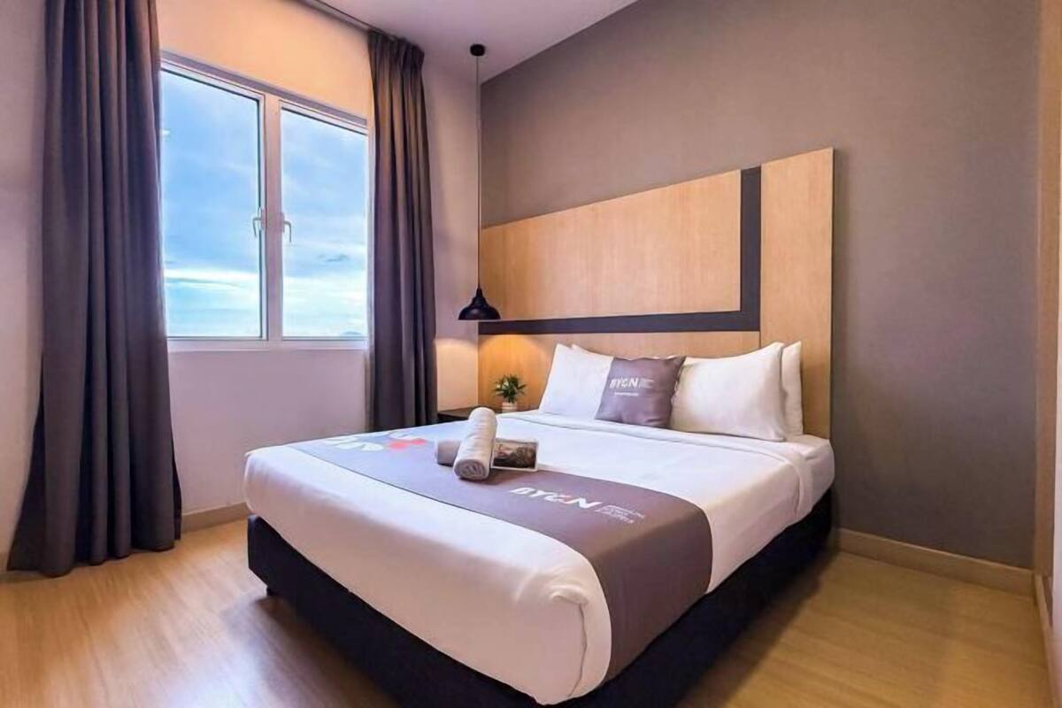 A hotel room with a bed, curtains, a window, and a wall-mounted lamp.