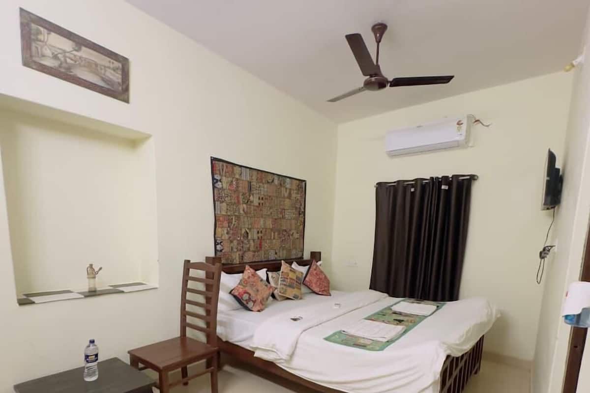 A bedroom with a bed, a chair, a small table, a ceiling fan, a wall-mounted air conditioner, and a pair of slippers on the floor.