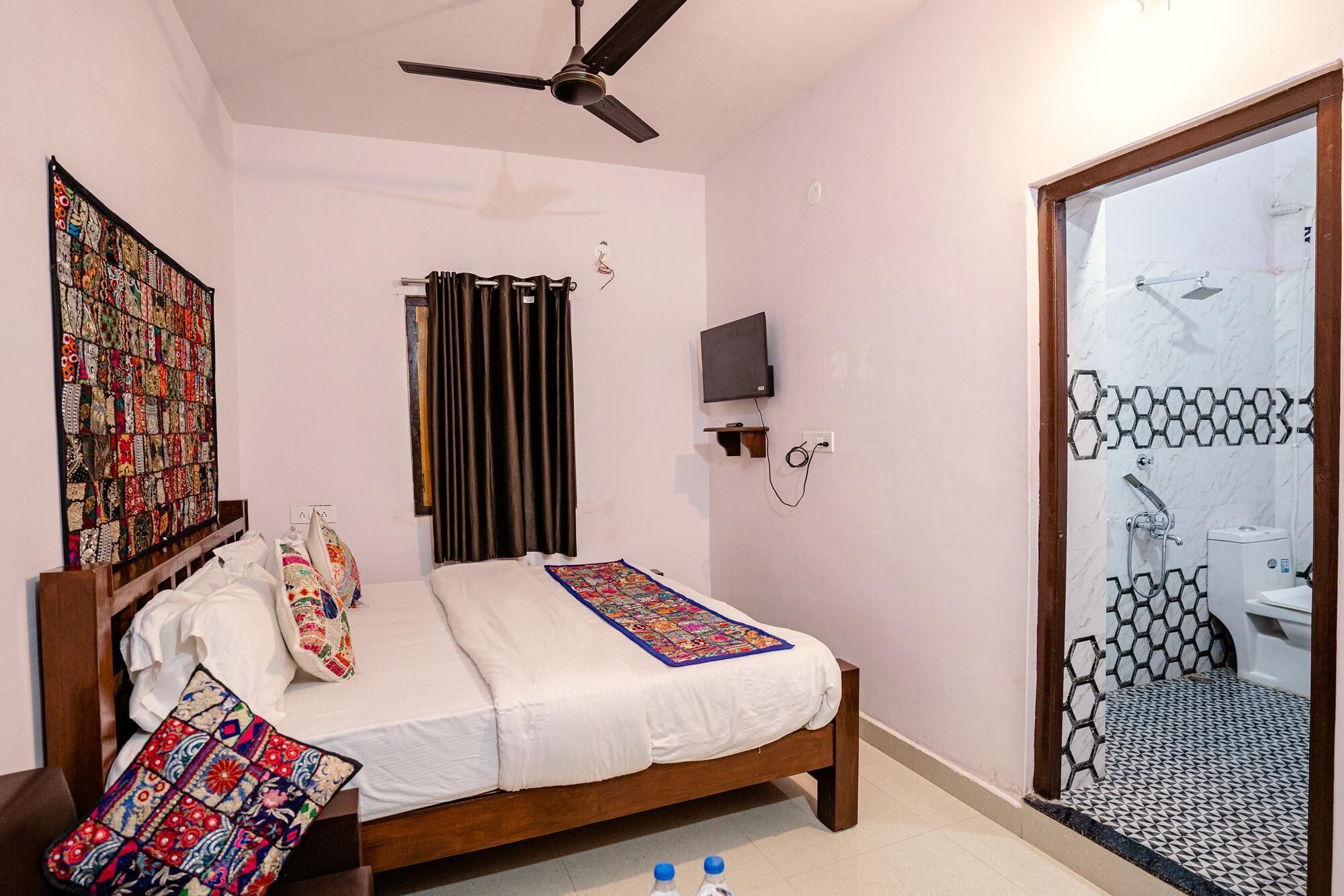 A bedroom with a bed, a ceiling fan, a TV mounted on the wall, and a bathroom with a shower and toilet visible through a glass door.