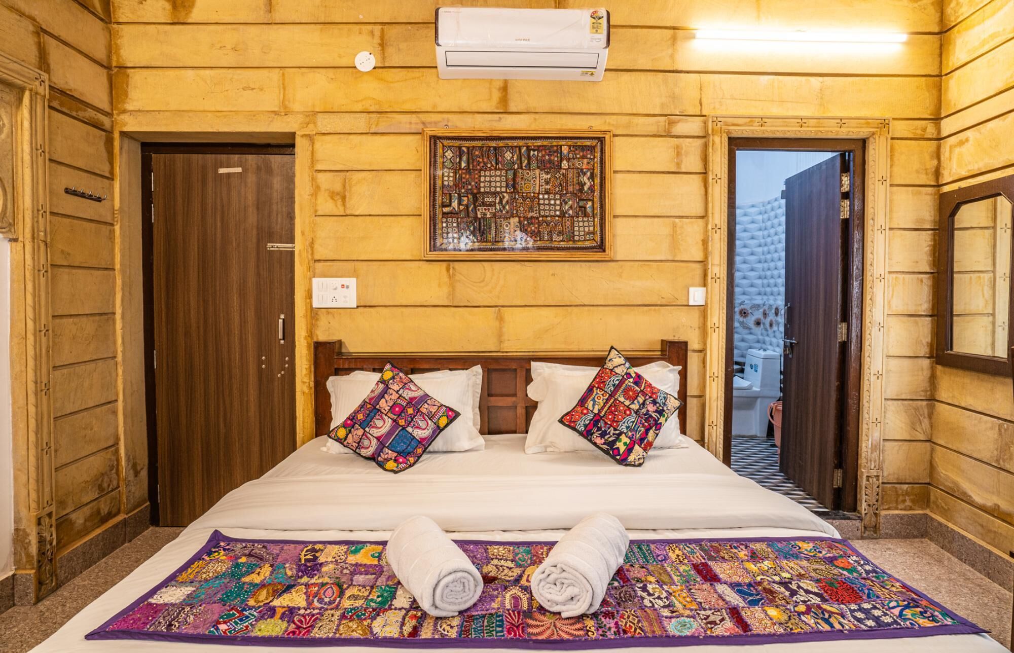 A room with a wooden wall, a bed with colorful pillows, a quilt, and a wall-mounted air conditioner.
