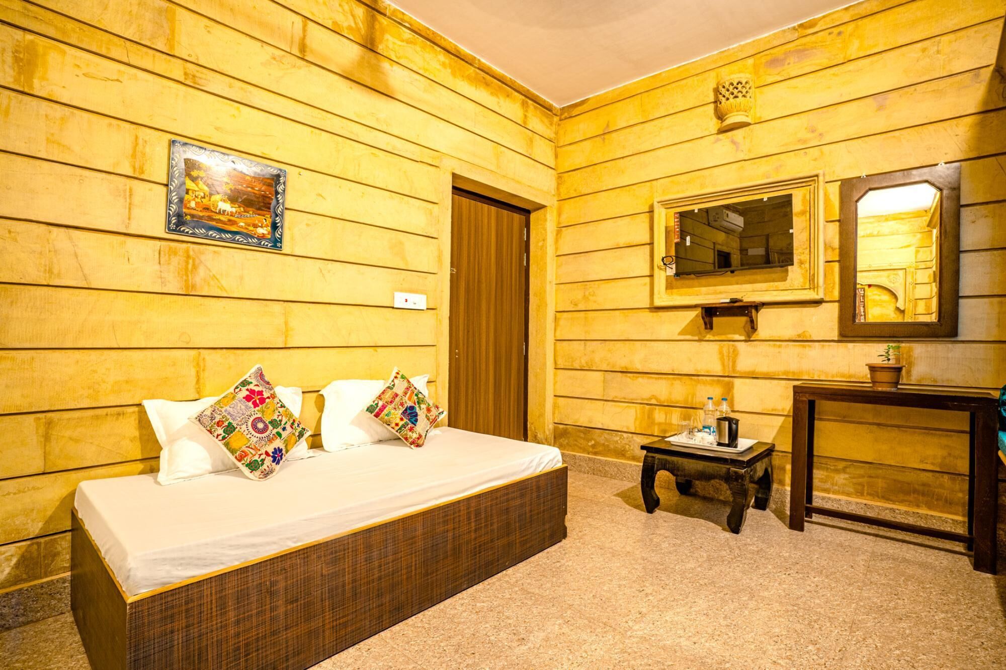 Una habitación con paredes de paneles de madera, una cama con ropa de cama blanca y almohadas de colores, una mesita de noche pequeña con una lámpara y un espejo sobre un tocador de madera.
