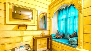 Um quarto com paredes de madeira, uma janela com cortinas azuis, uma pequena mesa com uma bandeja de garrafas de água e uma chaleira, e um espelho na parede.