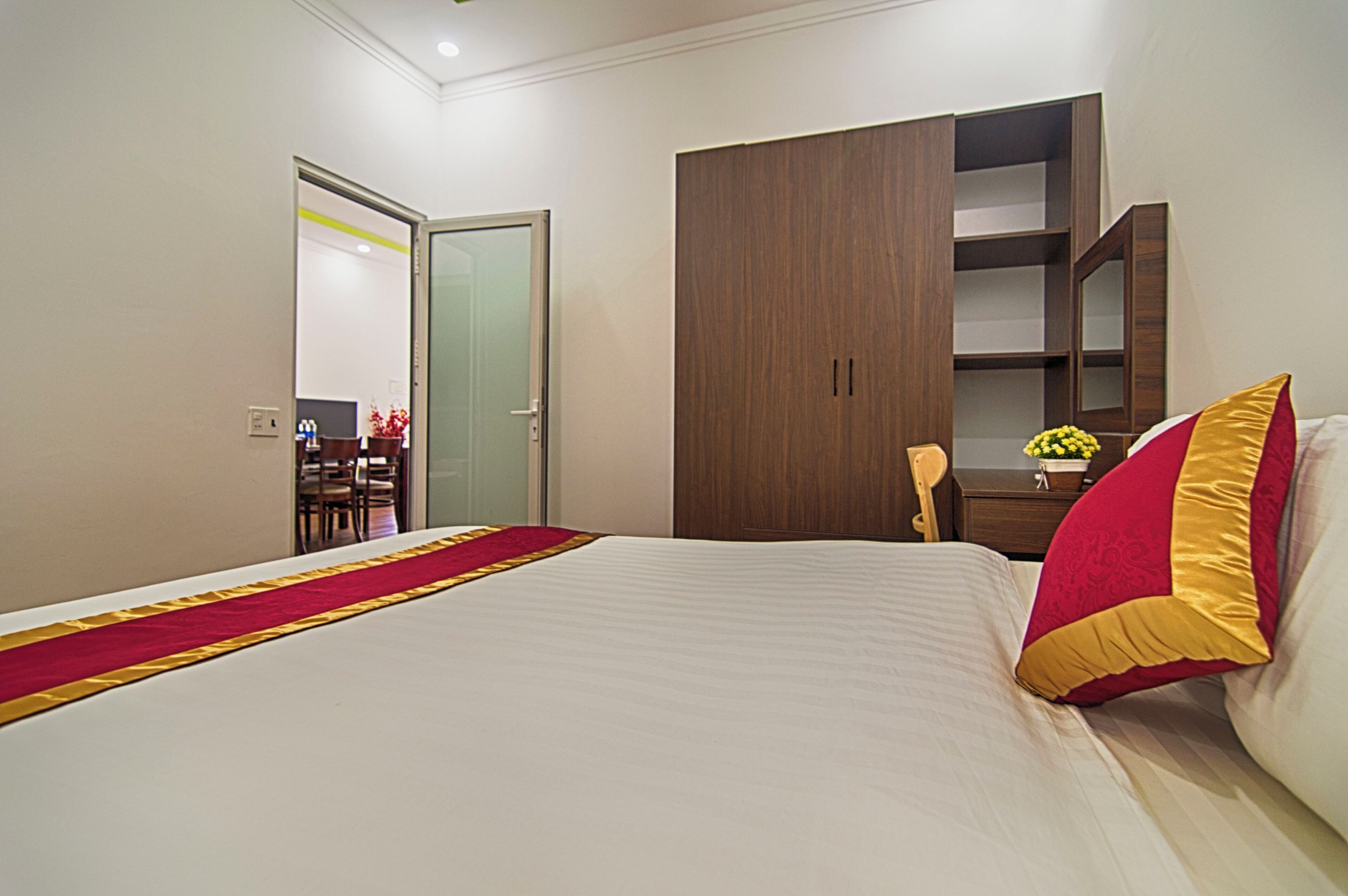 A hotel room with a large bed, a wooden wardrobe, a desk with a chair, and a small table with a vase of flowers.