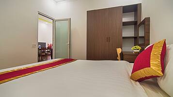 A hotel room with a large bed, a wooden wardrobe, a desk with a chair, and a small table with a vase of flowers.