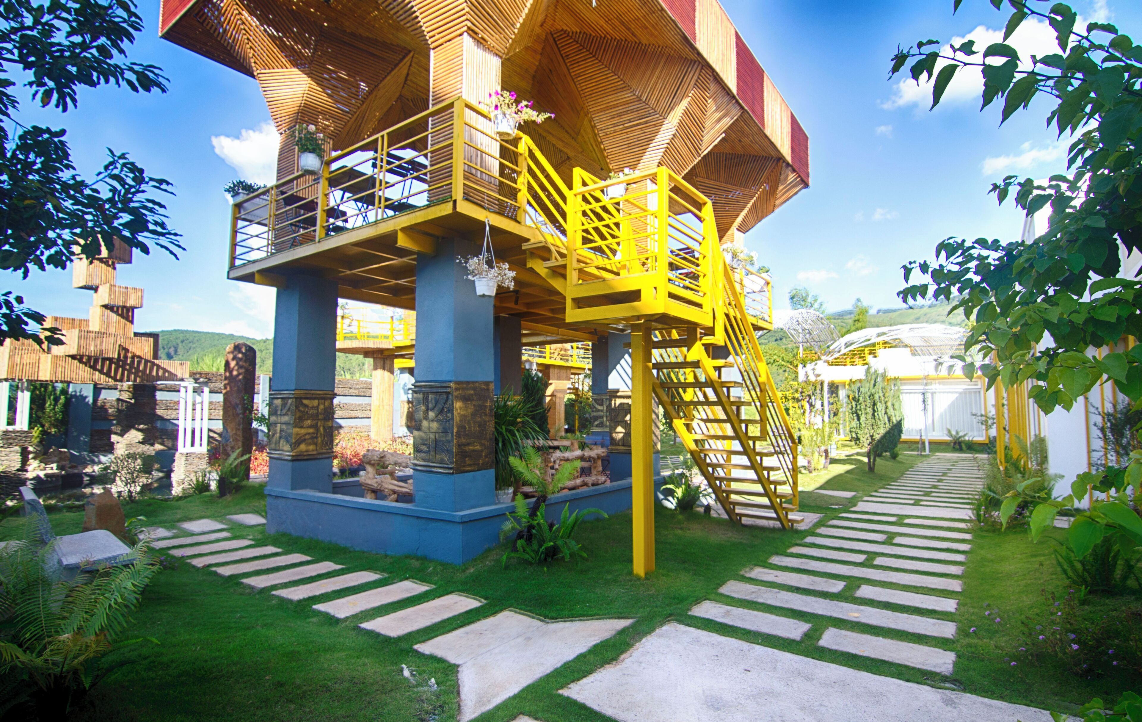 A multi-level wooden structure with a yellow staircase, surrounded by lush greenery and stone pathways.