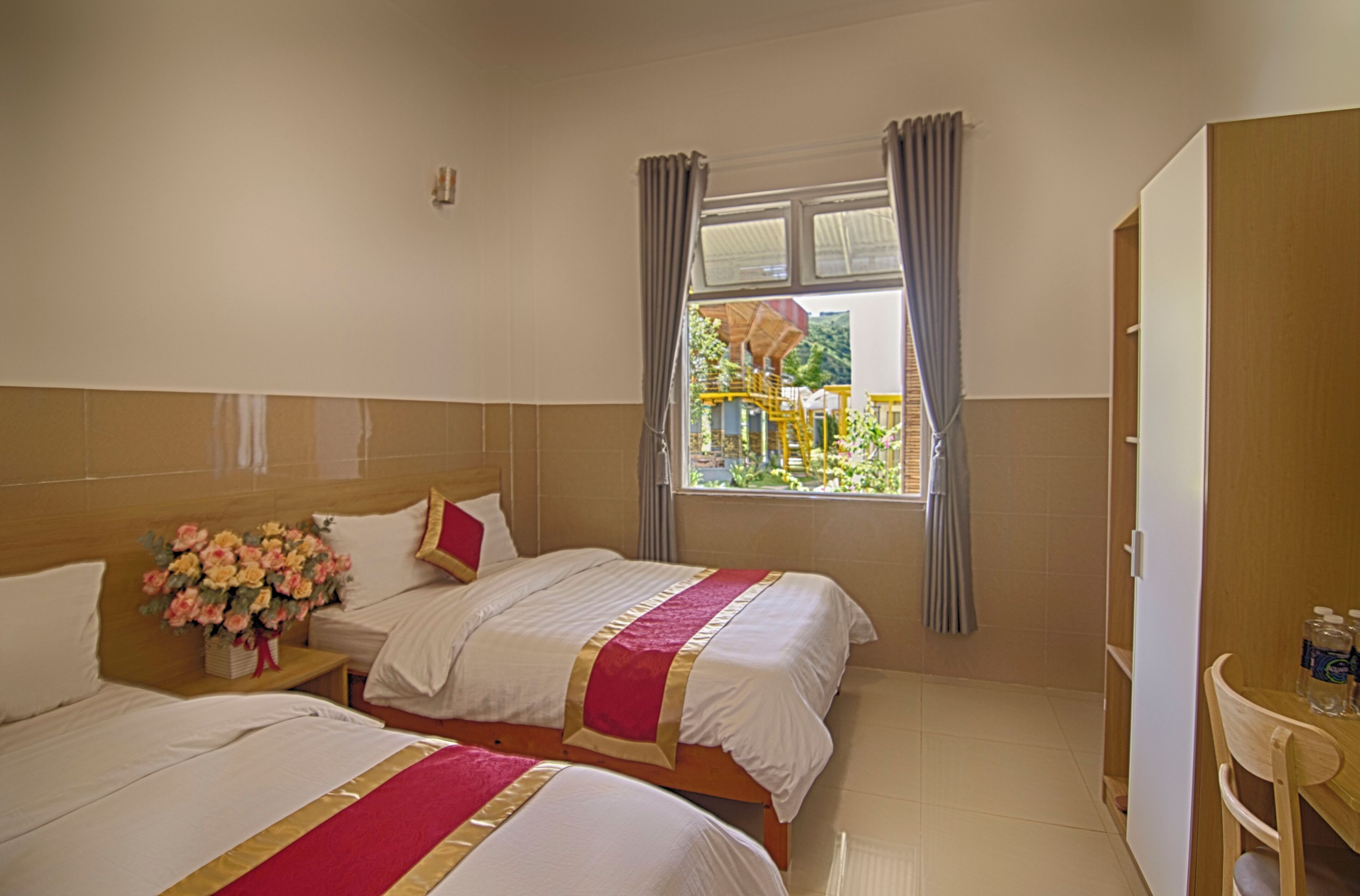 A hotel room with two beds, a window with curtains, a bedside table with a vase of flowers, and a wooden wardrobe.
