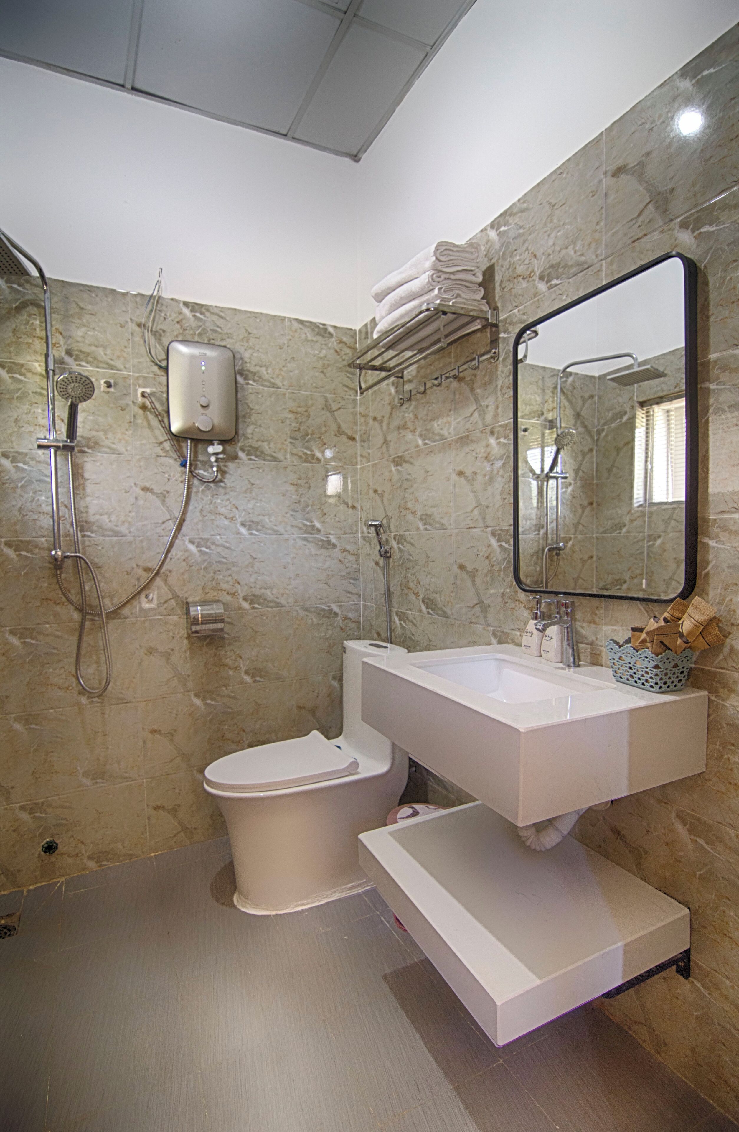 Un baño con pared de mármol, lavamanos blanco, inodoro, ducha y espejo.