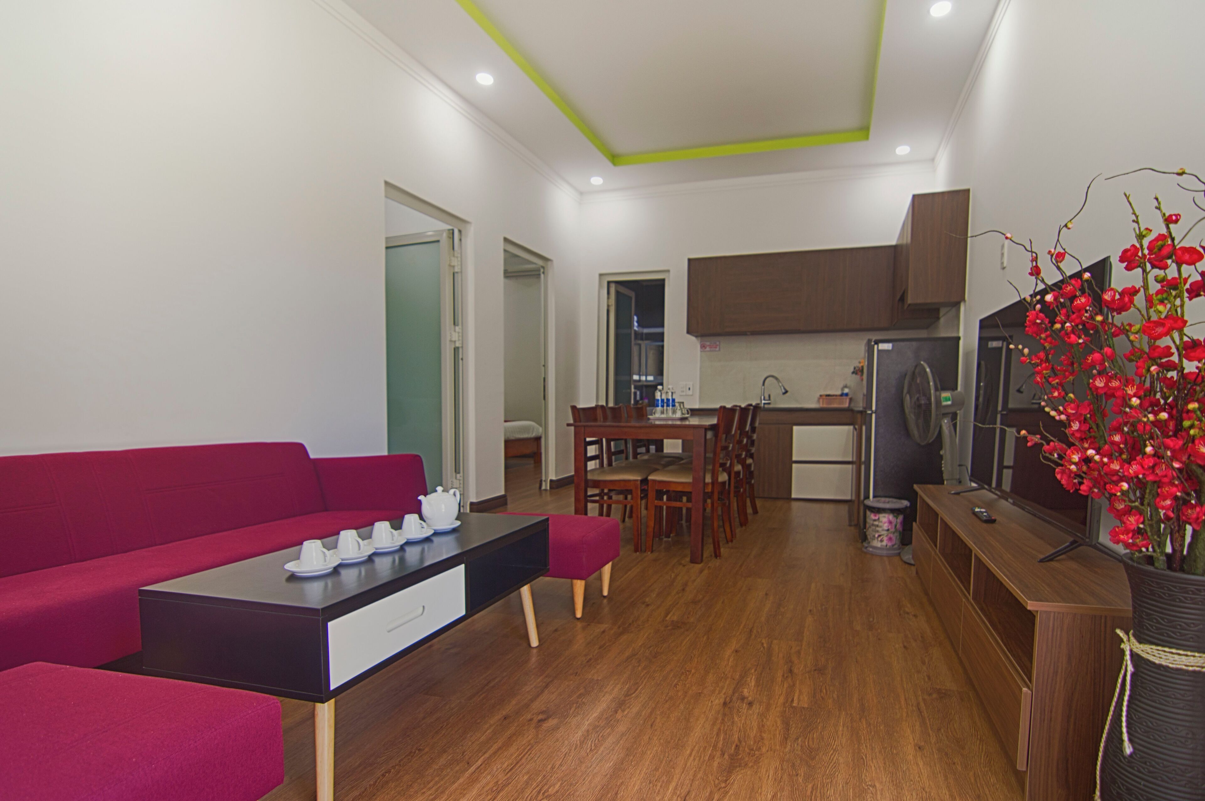 A modern living room with a red sofa, a dining table with chairs, a kitchen area with cabinets, and a decorative red flower arrangement.