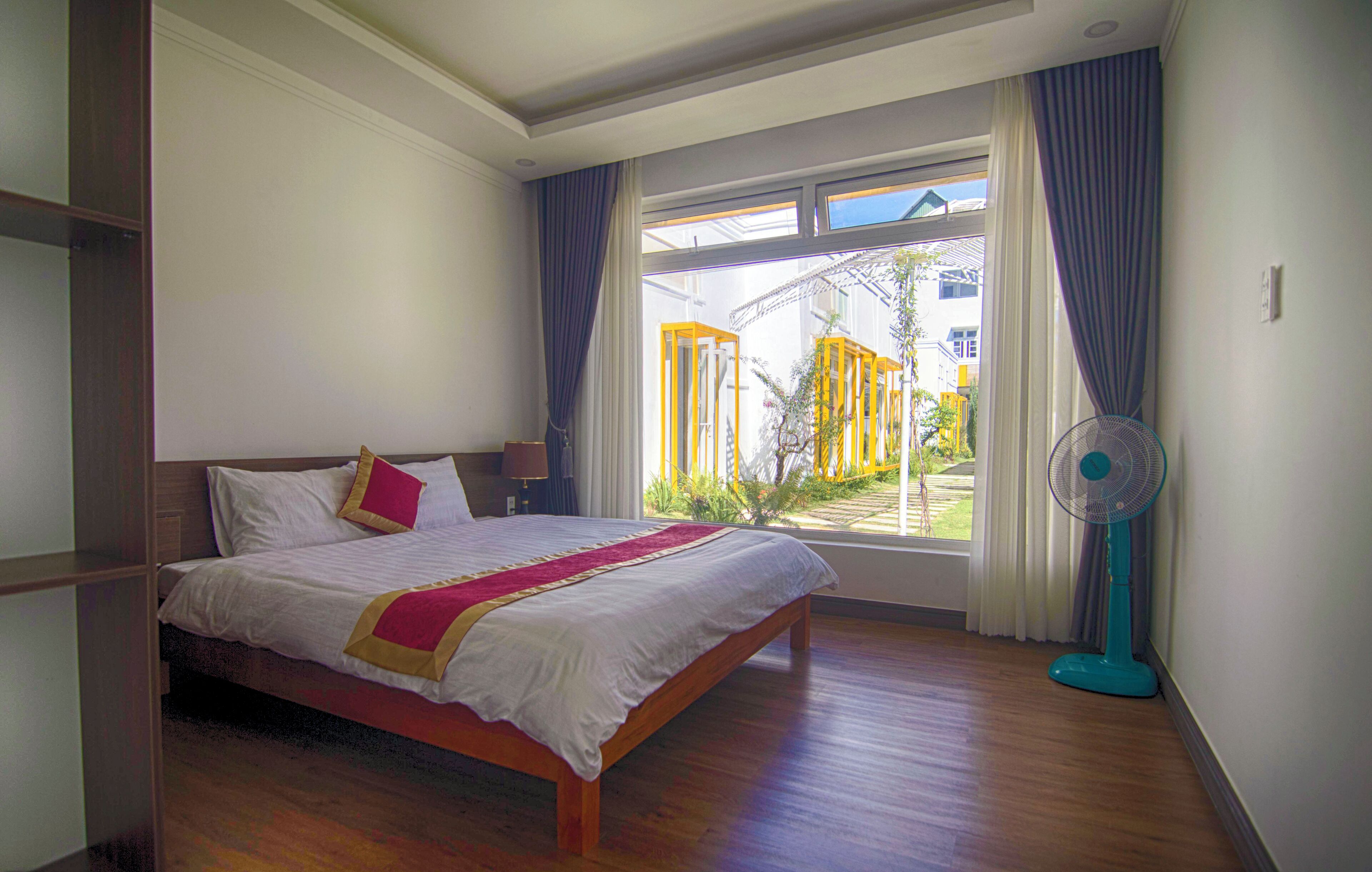 Un dormitorio con cama, ventana con cortinas, ventilador y vista a una estructura amarilla en el exterior.
