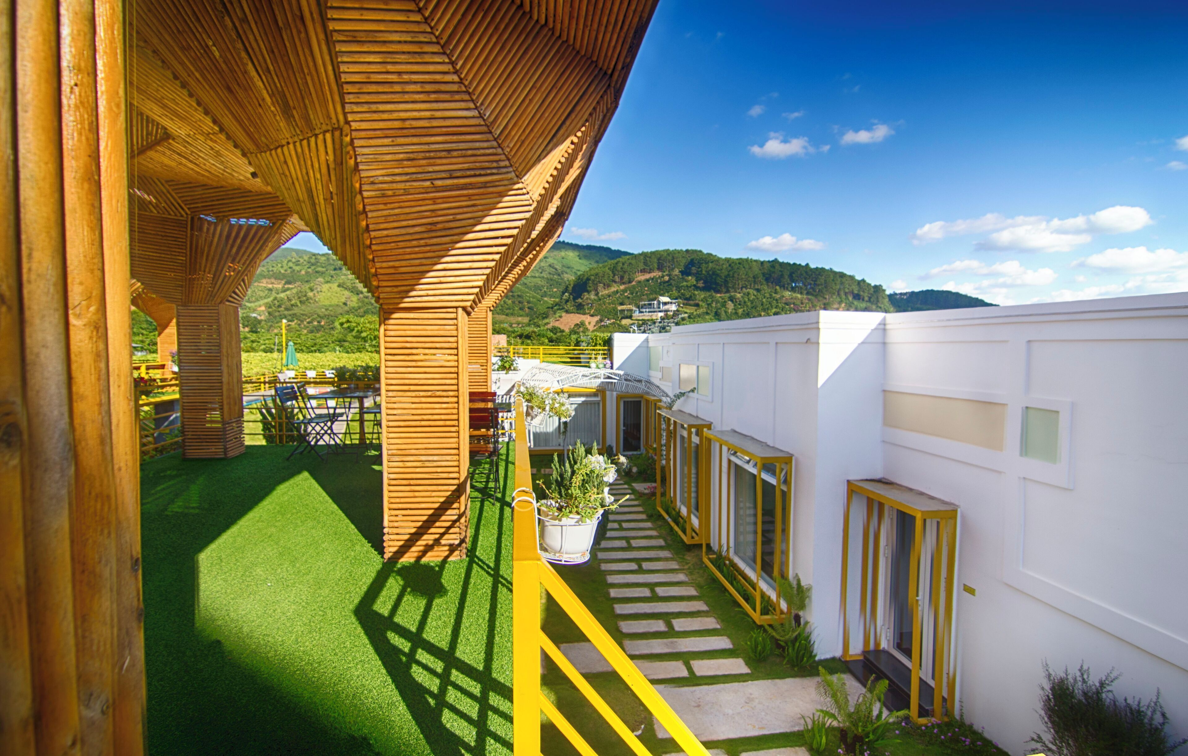 Un edificio moderno con estructura de madera, césped artificial verde y un sendero que lleva a un edificio blanco con barandilla amarilla.