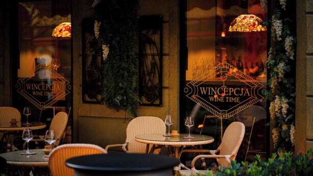 Un espace de restauration en plein air avec des chaises et des tables en osier, un mur décoratif agrémenté d’une plante suspendue et une enseigne indiquant : « WINCEPCA, l’heure du vin ».