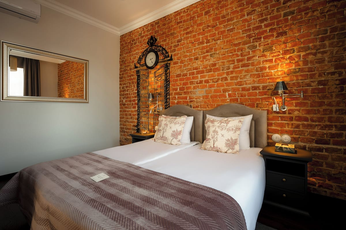 A hotel room with a brick wall, a large bed with a striped bedspread, two matching pillows, a nightstand with a lamp, and a mirror on the wall.
