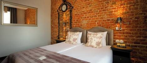 A hotel room with a brick wall, a large bed with a striped bedspread, two matching pillows, a nightstand with a lamp, and a mirror on the wall.