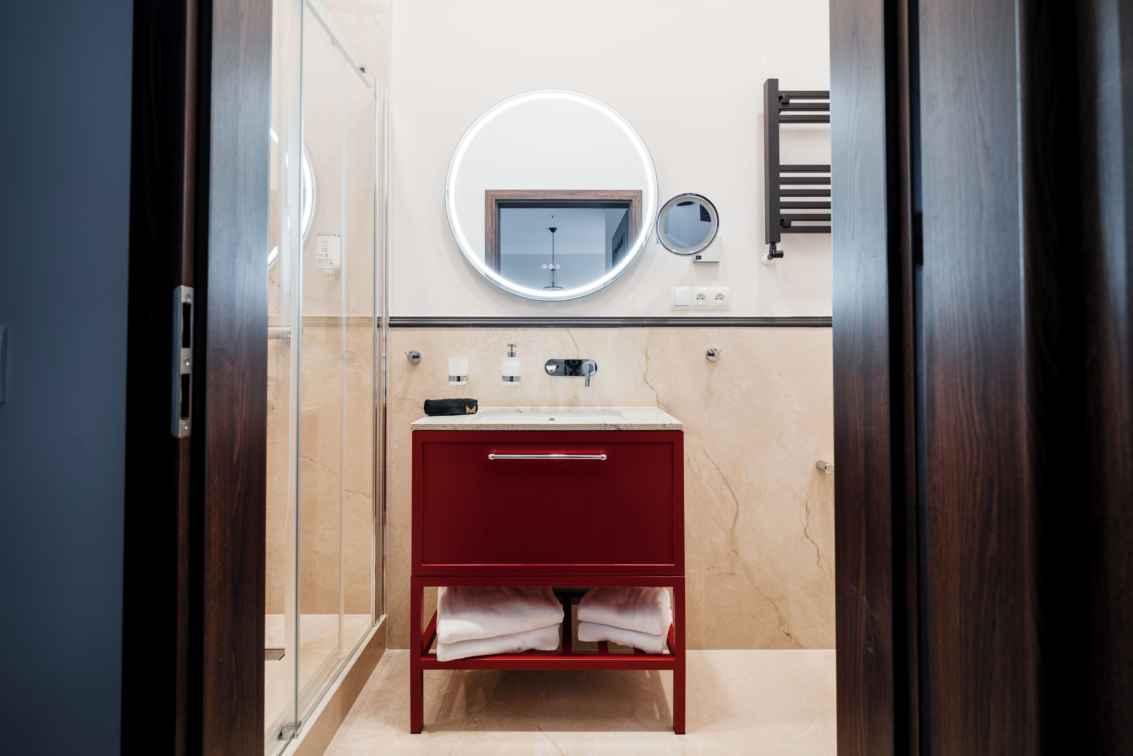 Un baño con cabina de ducha de vidrio, un mueble de tocador rojo con encimera de mármol, un espejo redondo y un perchero para toallas.