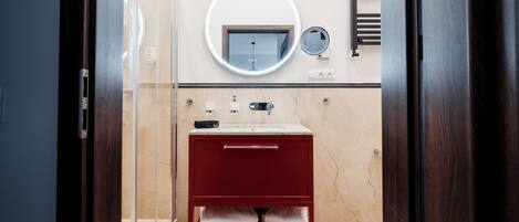 Un baño con cabina de ducha de vidrio, un mueble de tocador rojo con encimera de mármol, un espejo redondo y un perchero para toallas.