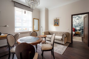 Sala de estar com mesa de madeira redonda, sofá bege e uma janela grande com persianas.