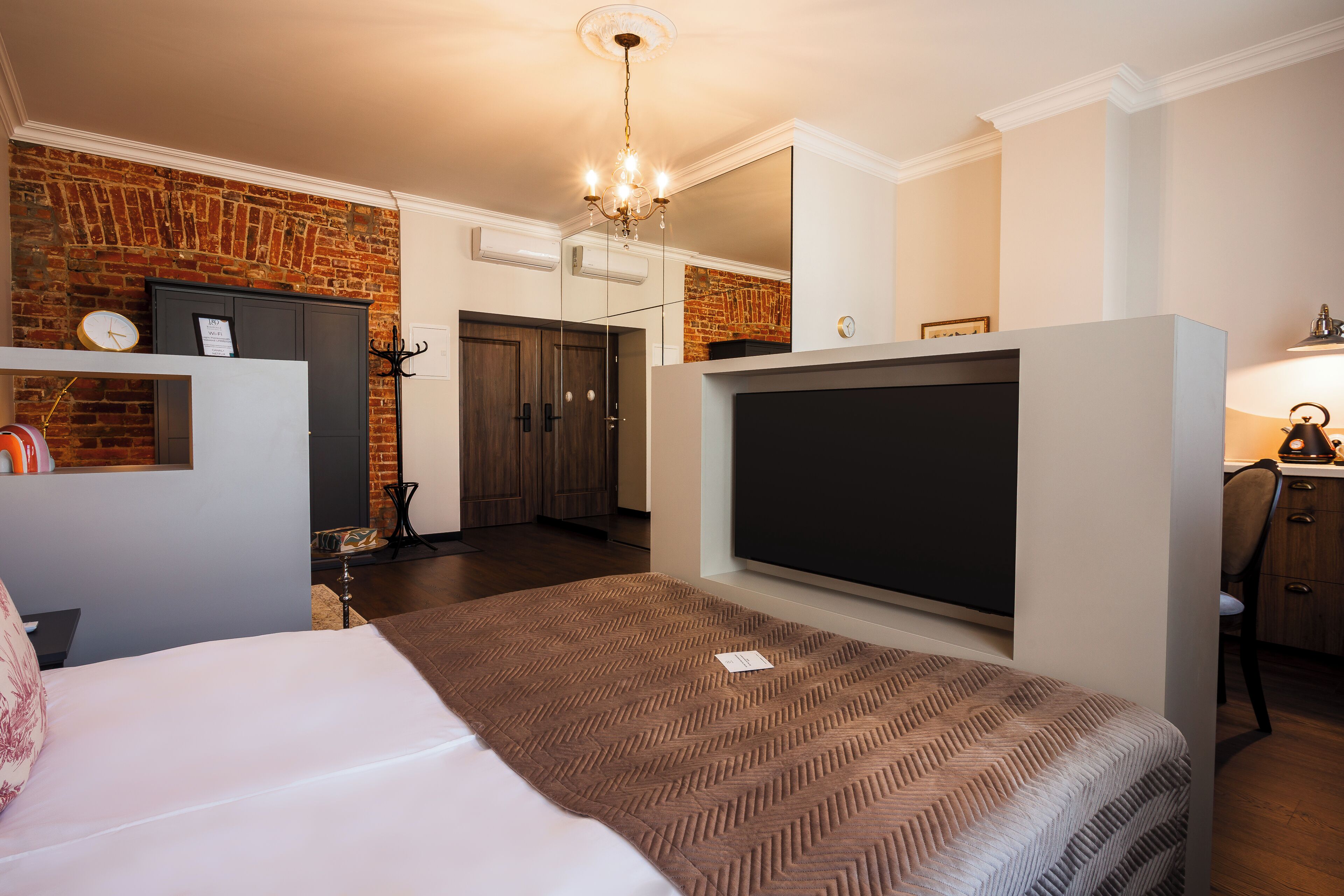 A modern hotel room with a brick wall, a large bed with a patterned bedspread, a flat-screen TV, a chandelier, and a desk with a chair.