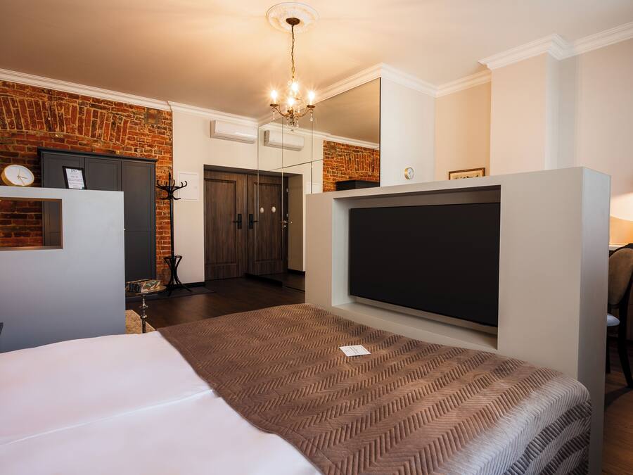 A modern hotel room with a brick wall, a large bed with a patterned bedspread, a flat-screen TV, a chandelier, and a desk with a chair.