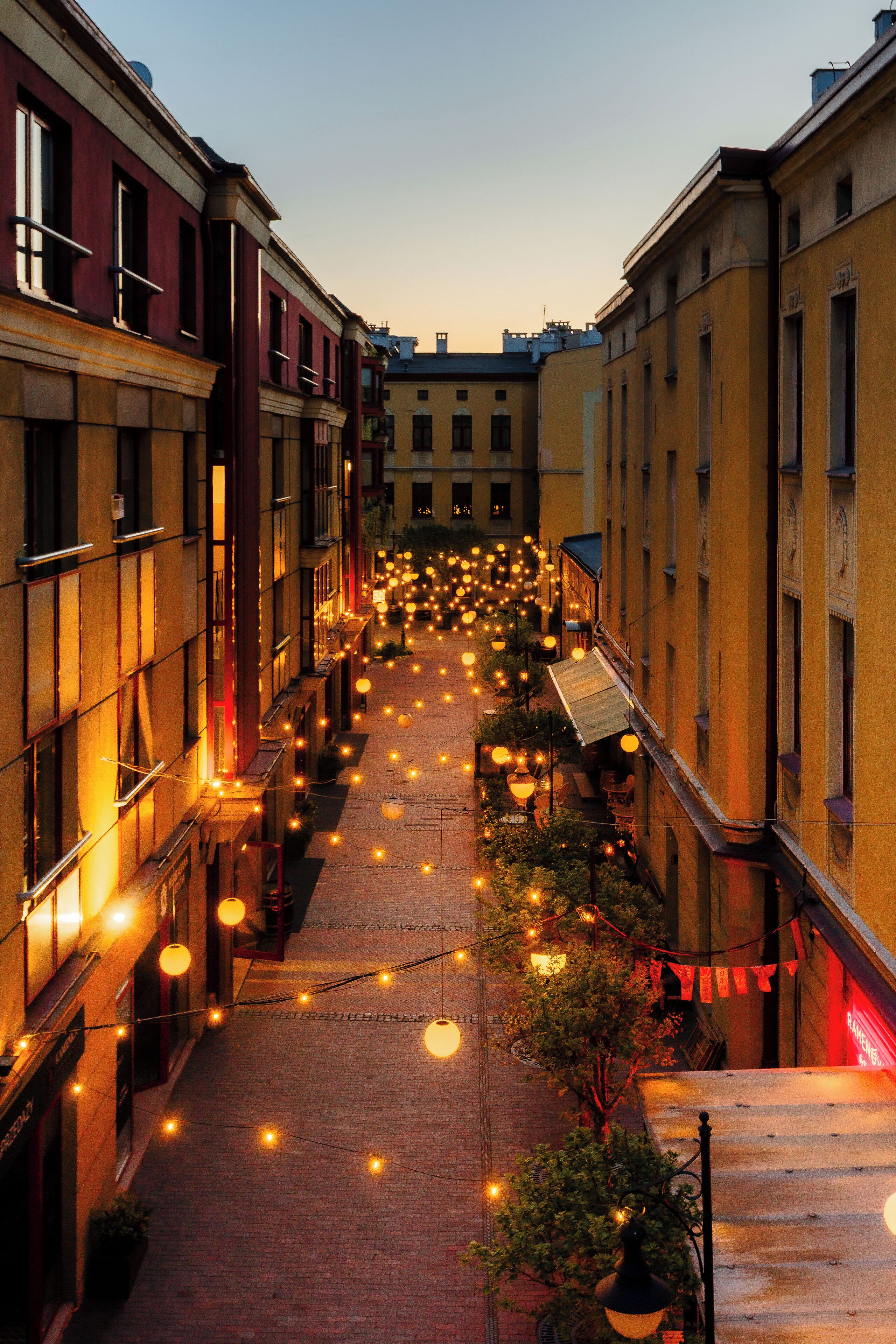 A narrow street lined with buildings, decorated with string lights and lanterns, suggesting a festive atmosphere.