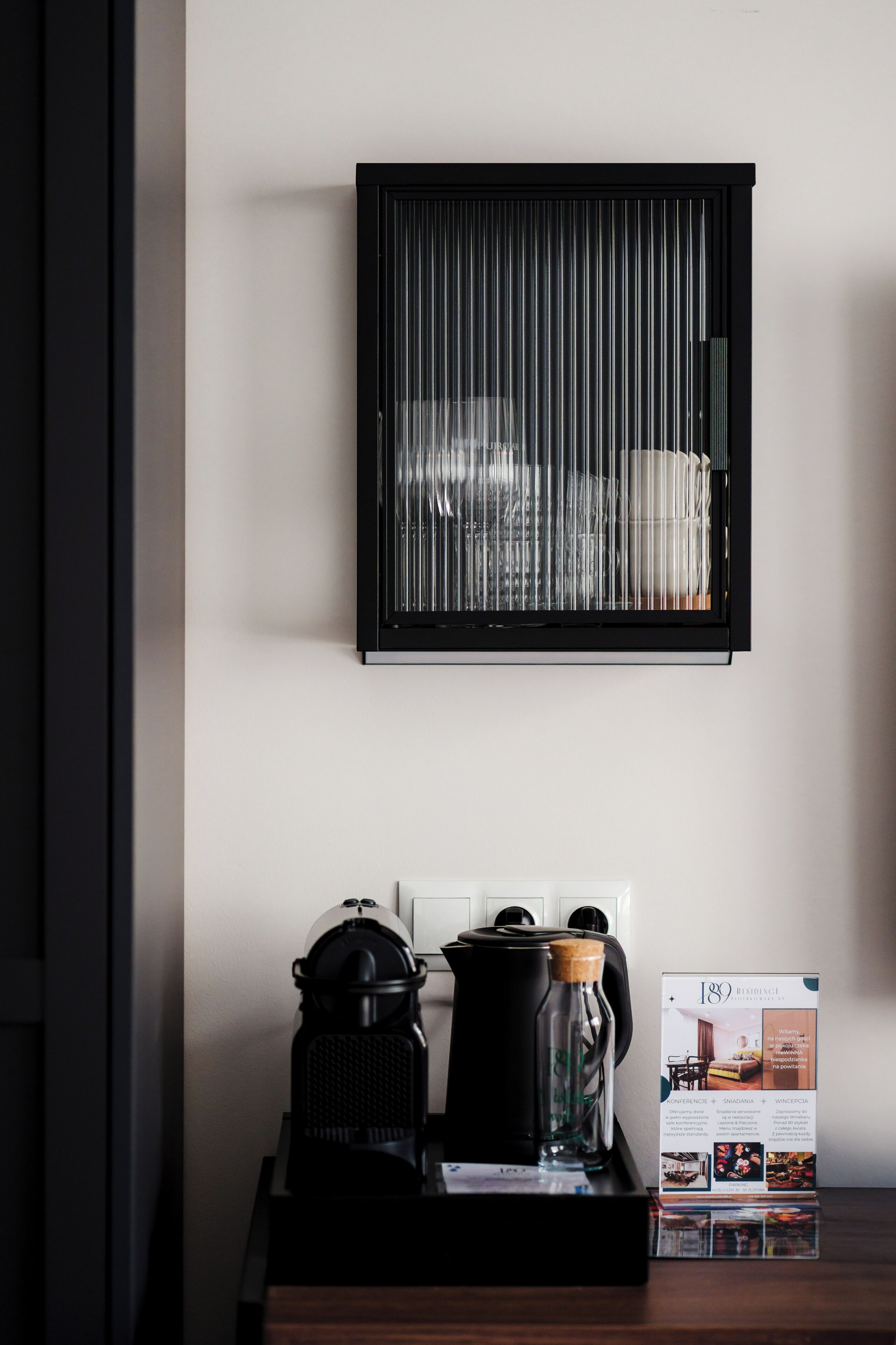Una ventana con marco negro y persianas verticales, una tetera negra, una jarra de vidrio y un folleto sobre una mesa de madera.