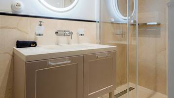 Un baño moderno con ducha de vidrio, un espejo redondo con iluminación integrada, encimera de mármol color beige y un perchero.