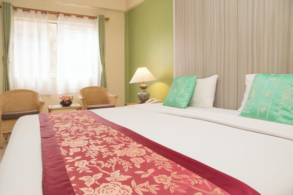 Une chambre d’hôtel avec un grand lit, deux fauteuils, une table de chevet avec une lampe et une vue sur l’extérieur à travers des rideaux légers.