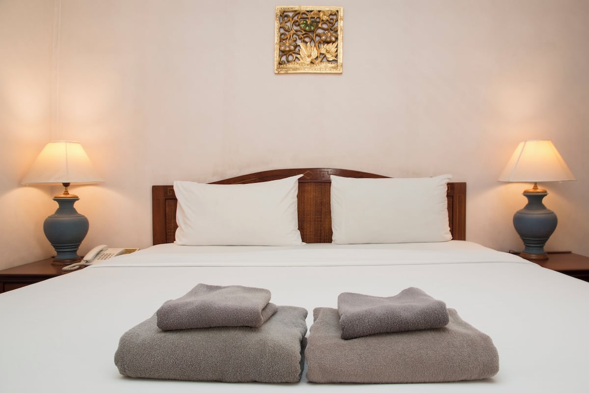 Un lit bien fait, avec du linge de lit blanc et deux oreillers, flanqué de deux tables de chevet sur lesquelles reposent des lampes, et un élément décoratif mural au-dessus du lit.