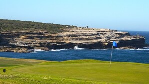 Un campo de golf costero con una bandera en el green, acantilados rocosos al fondo y un cielo despejado.