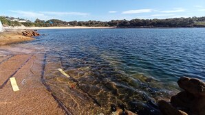 Una vista despejada de una playa con costa rocosa, agua tranquila y una línea de costa distante con edificios y árboles.