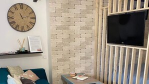 A living room with a teal sofa, a wooden TV stand, a brick-patterned wall, and a round wall clock. - Studio Apartment 'La Noisette' with Private Terrace, Private Garden and Wi-Fi (Equemauville)